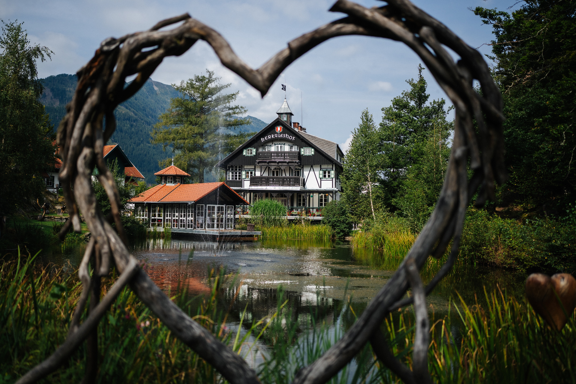 Blick durch einen Herzrahmen aus Ästen auf den Herkuleshof und den Teich